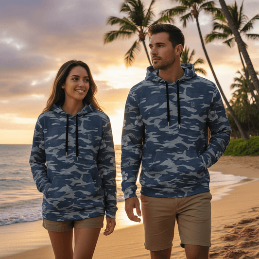 Couple wearing matching Pacific Warrior pullover hoodies with blue camo and hibiscus flowers walking on Hawaiian beach at sunset