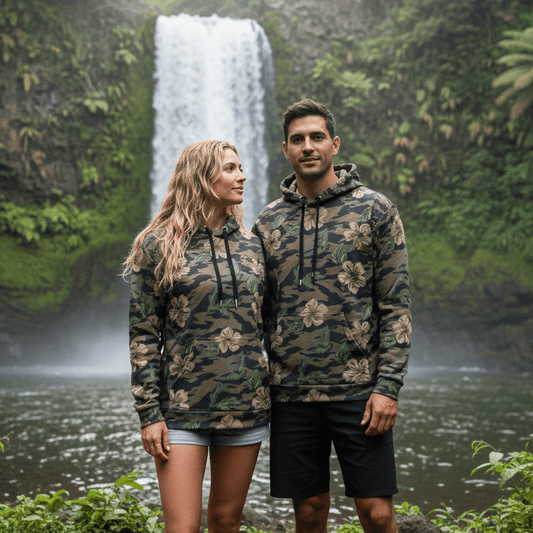 Couple wearing matching Island Guard pullover hoodies with military camo and tan hibiscus flowers at tropical waterfall