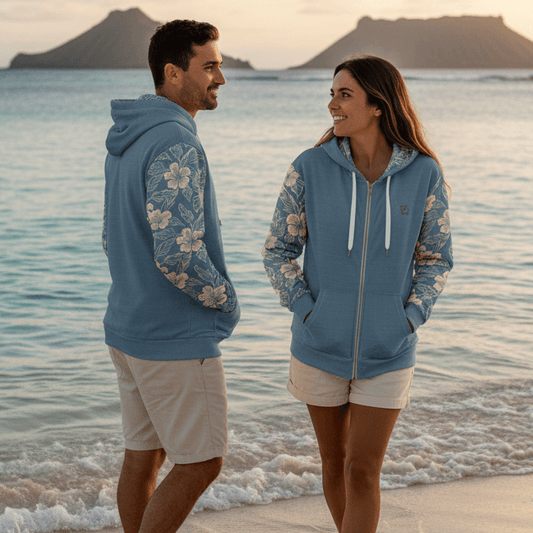 Man and woman wearing matching Kailua Opus dusty blue zip hoodies with cream hibiscus flowers walking along Lanikai Beach at sunset