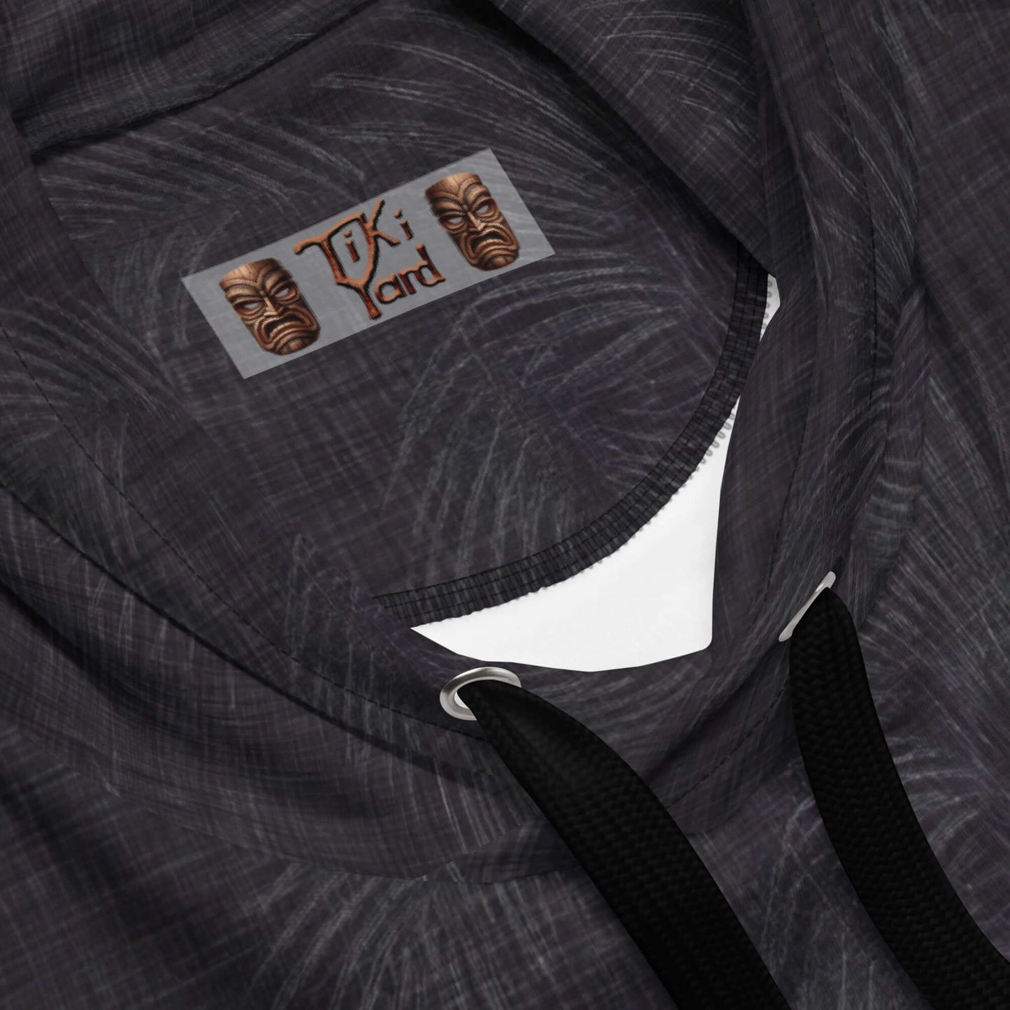 Close-up view of the Tiki Yard label on a black Hawaiian hoodie with intricate white pattern and black drawstrings.