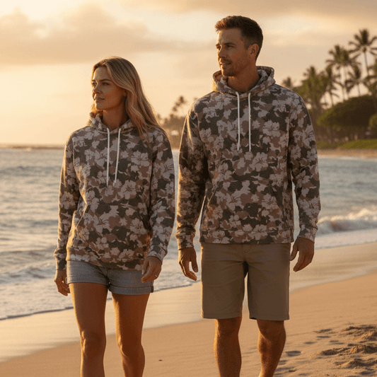 Couple wearing matching Beach Crusader pullover hoodies with camo and pink hibiscus flowers walking on Hawaiian beach at sunset