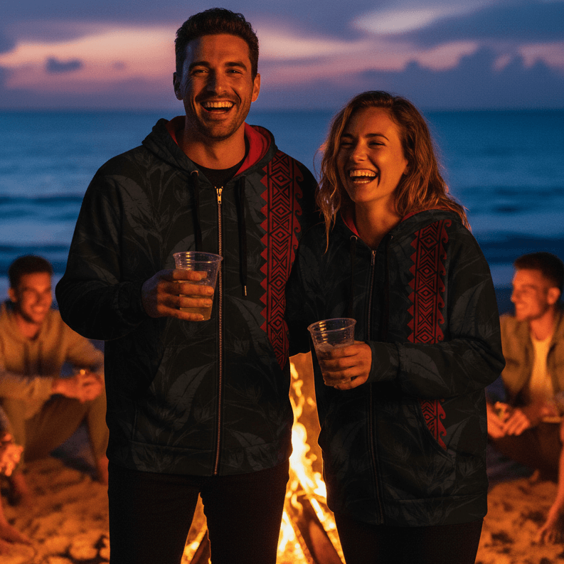 Man and woman wearing matching Po-Koa-o-ke-Kai black tropical leaf zip hoodies at evening beach bonfire with friends